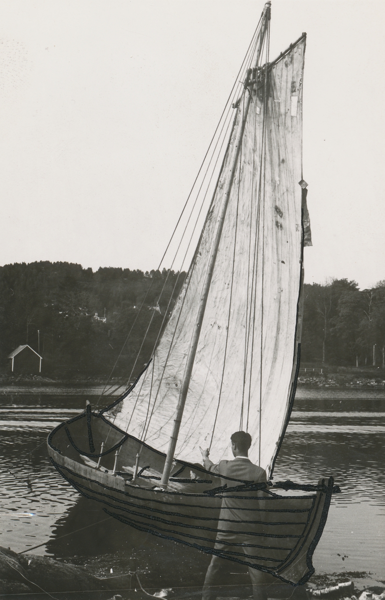 En sunnmørsåttring under seil i fjæra i Katavågen ved Sunnmøre Museum. En mann i dress står i fjæra og peiker på båten. En annen mann sitter oppi båten.