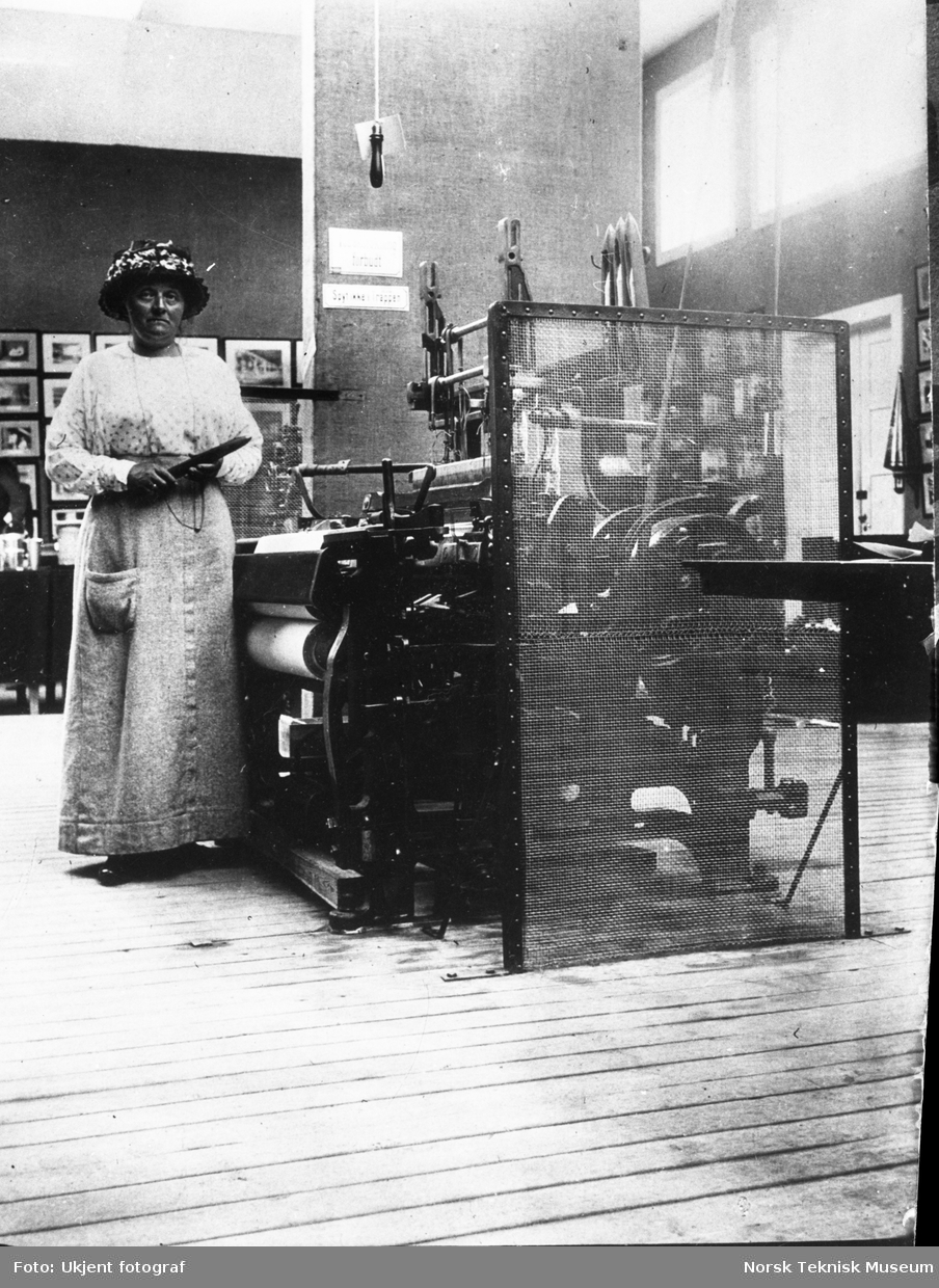 Betzy Kjelsberg ved en vevstol med beskyttelsesgitter, med skyttel i hånden og iført forkle (arbeidstøy). I bakgrunnen skilt der det står "Spyt ikke i trappen". Fra Arbeiderbeskyttelsesutstillingen i Kristiania i 1914.