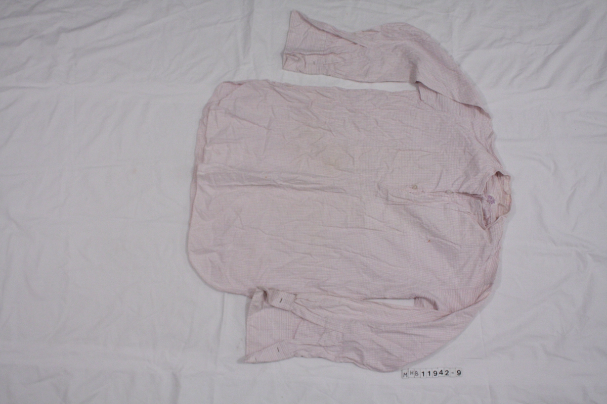 Skjorte uten krage. Hvit med lilla rutemønster. Kun åpning i øvre del av skjorte, lukkes med to knapper.