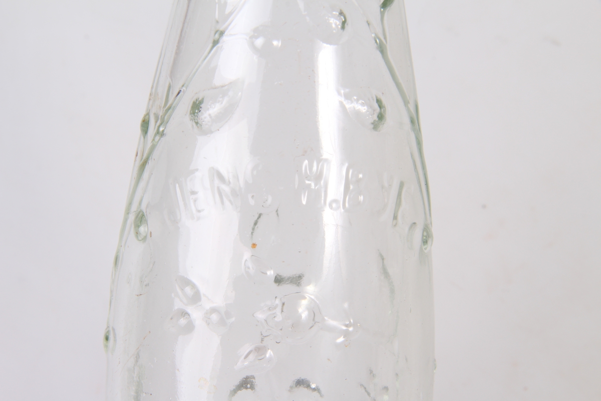 Smal høy flaske av klart glass. Langsgående dekor av bladranker støpt inn. "Jens H. Bye", støpt inn i øverste del.