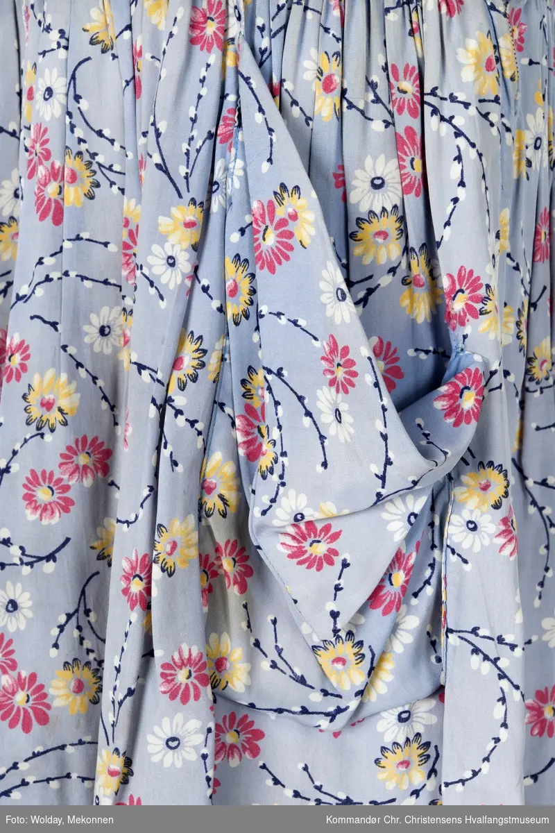 Trekvartlang kjole. Mønster er gråblå bunn med rosa, gule og hvite blomster, samt "puselabber".  Korte ermer. Glidelås av metall i siden.