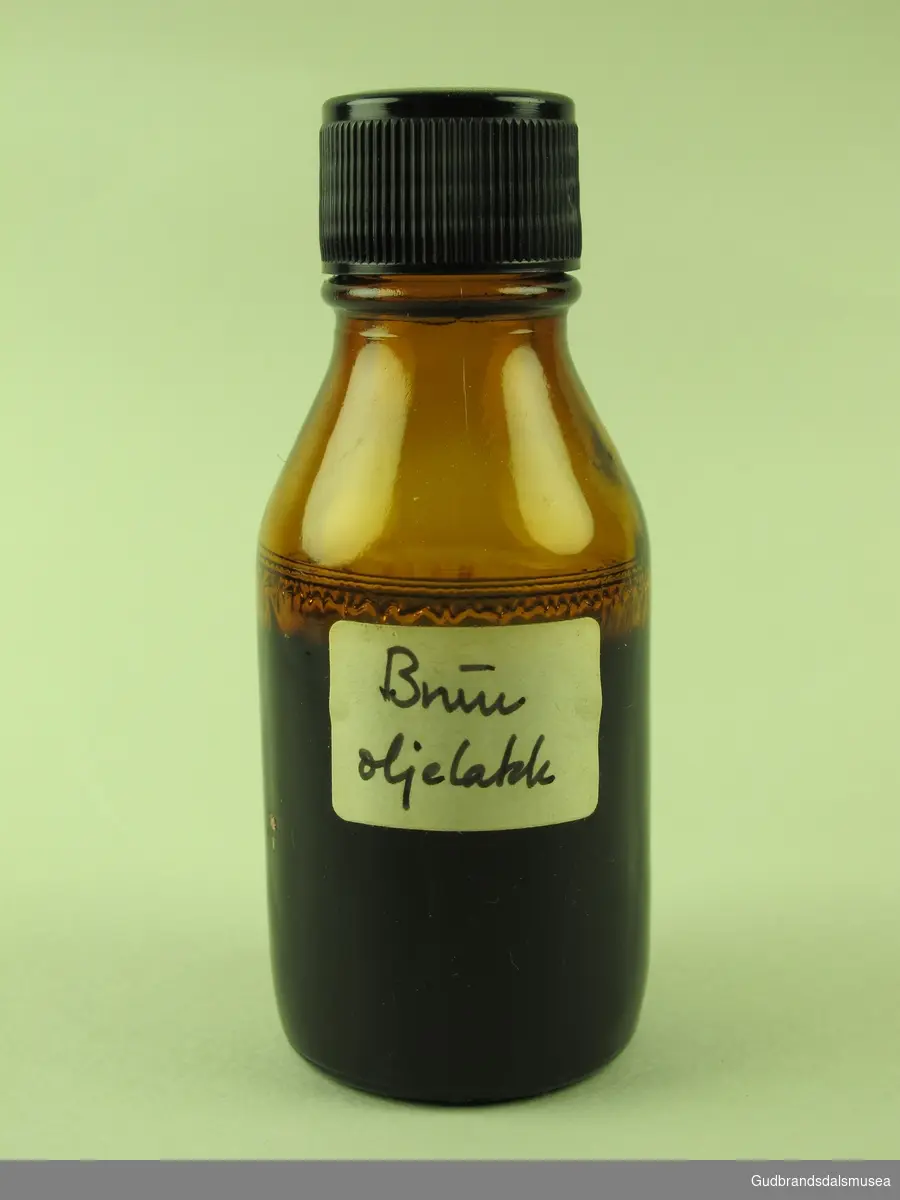 Brun rund glassflaske med sort plastkork. Hvit klistrelapp som etikett med blyantskrevet tekst.