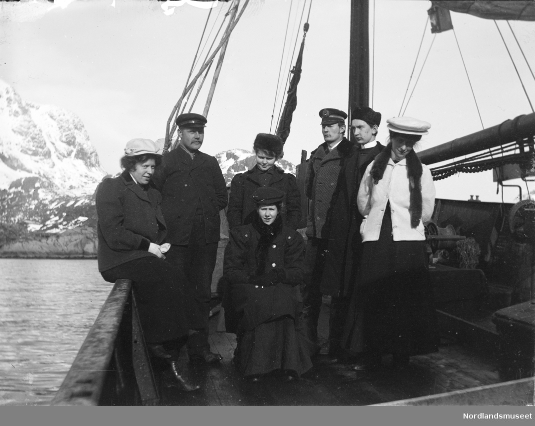 Oppsynsskøyte "Harriet", Skipper Johan Hass t.v., sammen med fire kvinner og to menn ombord i båten. Ved masten: A. Anderssen-Strand. Alle personene på bildet er finkledde. I bakgrunnen fjell. Negativen er en glassnegativ.