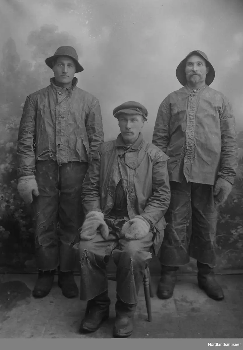 Portrett. 3 menn i 20-30-åra, i arbeidsklær/oljehyre. En sitter, to står bak. En med sydvest, en med hatt og en med lue. Alle med votter.