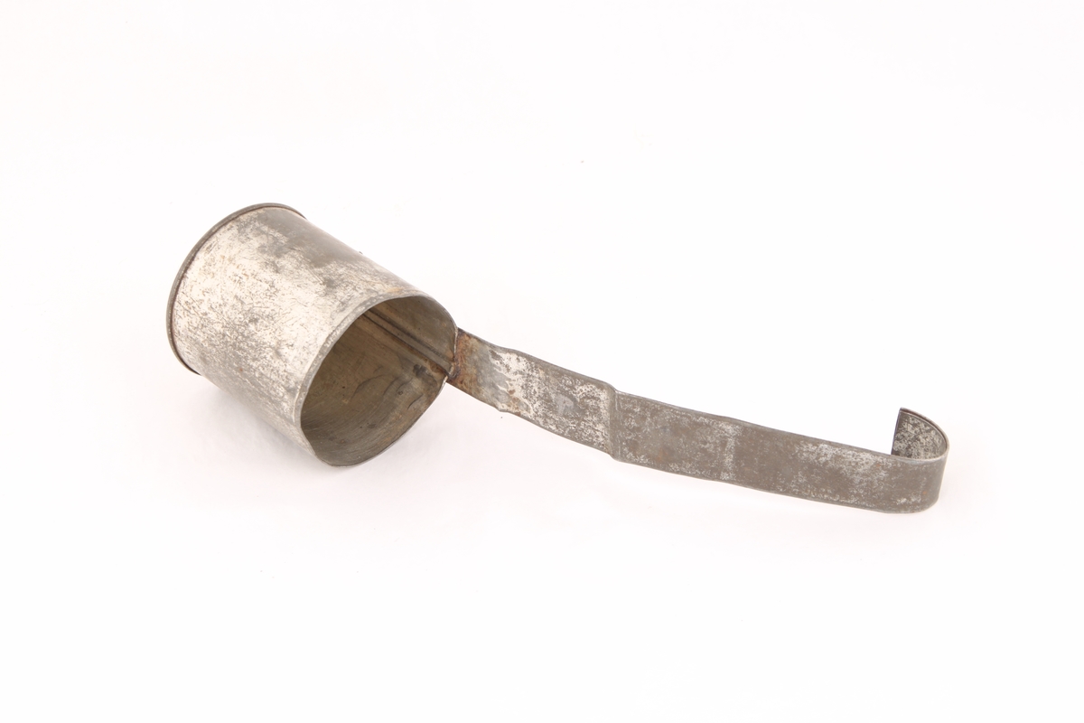 En liten øse formet som en kopp i metall med lang flat hank formet av brettet metall. Hanken er loddet eller sveiset fast til koppen.