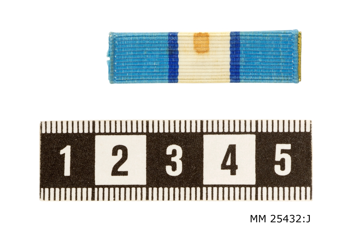 Släpspänne i sidenrips med nål på baksidan att fästa i uniformen. Uppspänt på mässingsplatta. Består av ljusblå fält vid kanterna och i mitten ett vitt fält. Dessa är separerade av mörkblå linjer. En rödbrun fläck finns på det vita fältet.