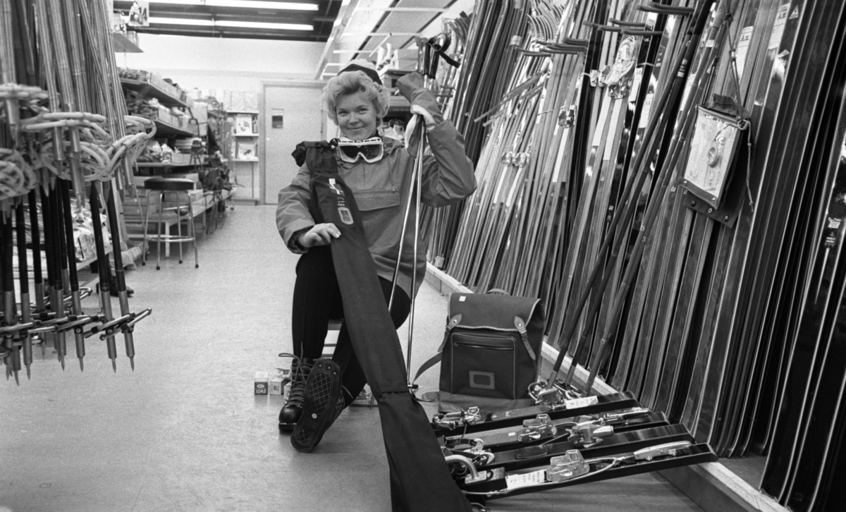 Fjälldags 16 februari 1966

I bildens förgrund sitter en vinterklädd kvinna med pjäxor på fötterna. Hon håller i ett par skidstavar med vänstra handen. Över högra axeln har hon lagt ett par skidor i ett skidfodral. Hon befinner sig inne i en affär som säljer sportutrustning. Till höger om henne står massor med skidor. En ryggsäck står på golvet. Till vänster syns skidstavar hänga. I bakgrunden syns lagerhyllor, en pall samt en ytterdörr.