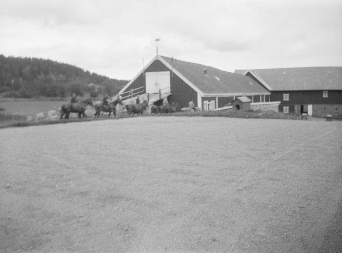 Gårdsplass og driftsbygning med låvebru på Linderud Gård. Fem mennesker til hest, samt noen flere hester uten ryttere.
