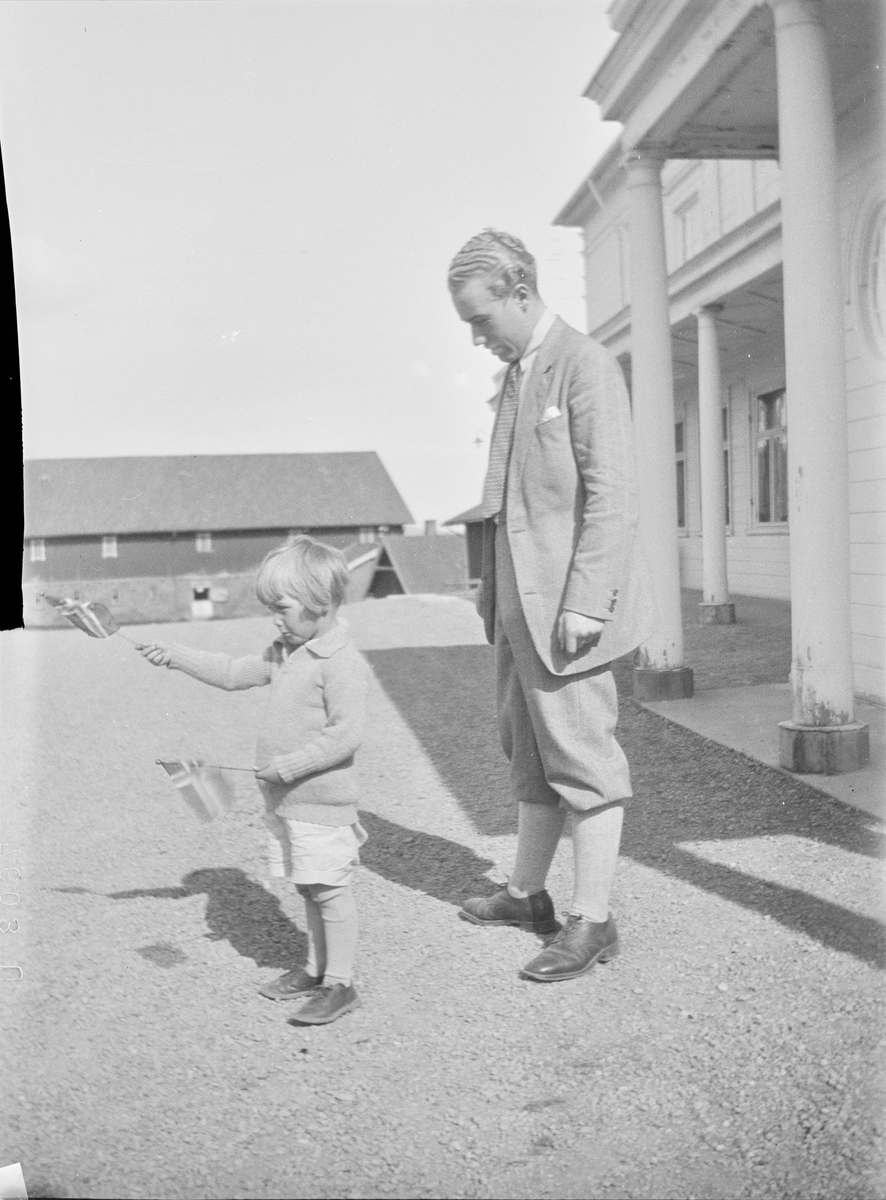 Iacob Ihlen Mathiesen står på gårdsplassen utenfor svalgangen til hovedhuset på Linderud Gård sammen med et barn som vifter med ett norsk flagg i hver hånd.