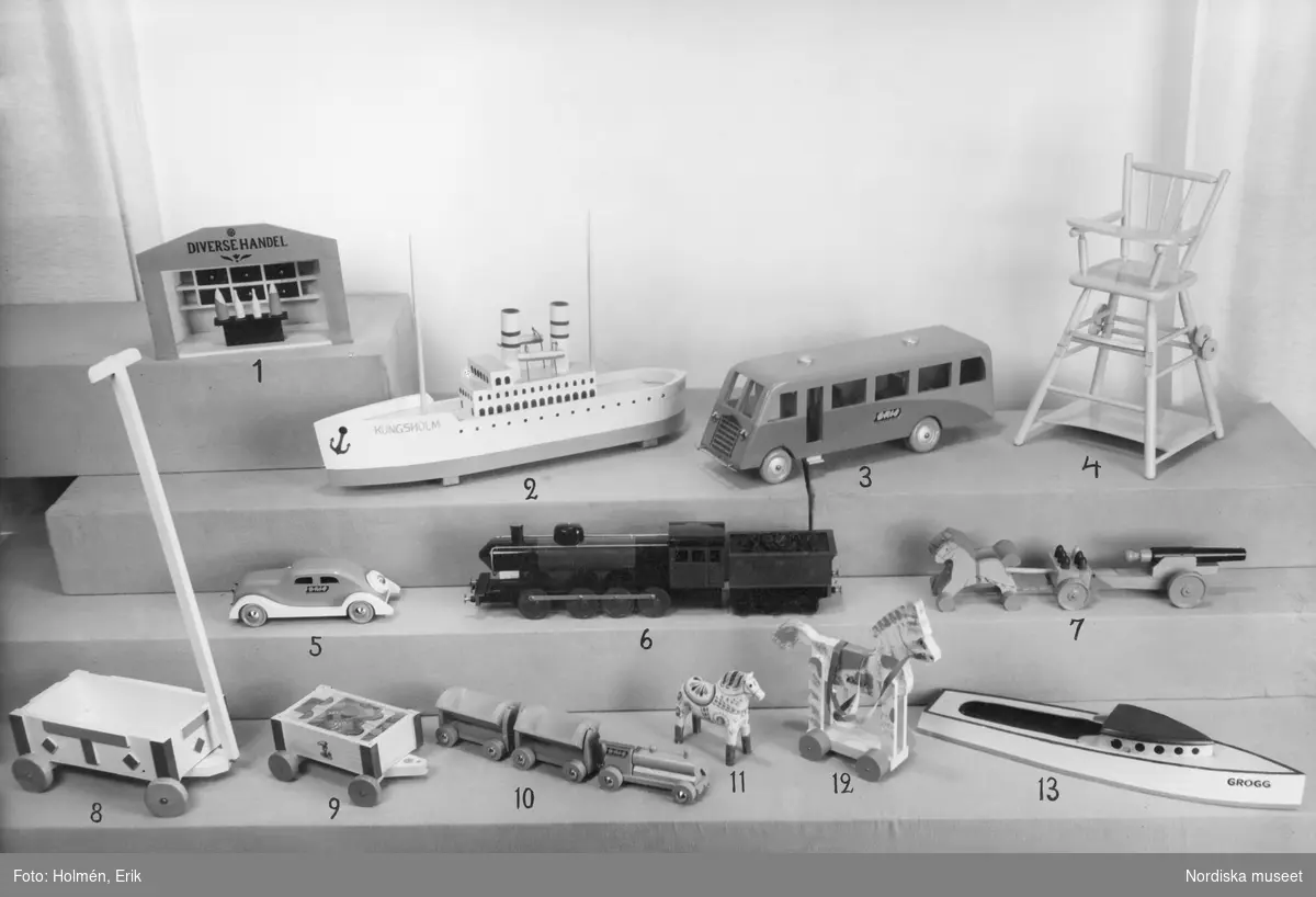 På hyllplan står olika leksaker uppradade tillsammans. Båtar, bil, buss, tåg, en diversehandel och annat.
