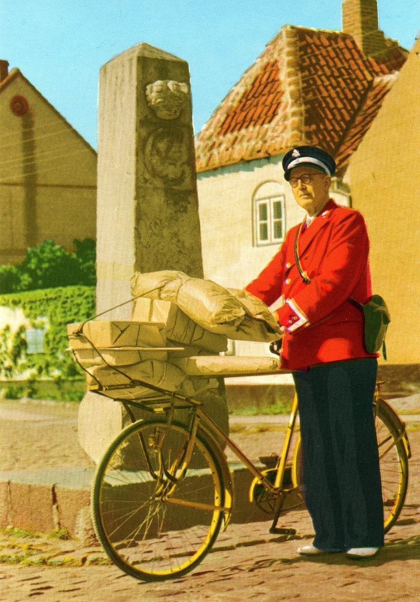 Vykort med motiv från ett fotografi av en dansk brevbärare med cykel och många paket invid en gammal milsten.