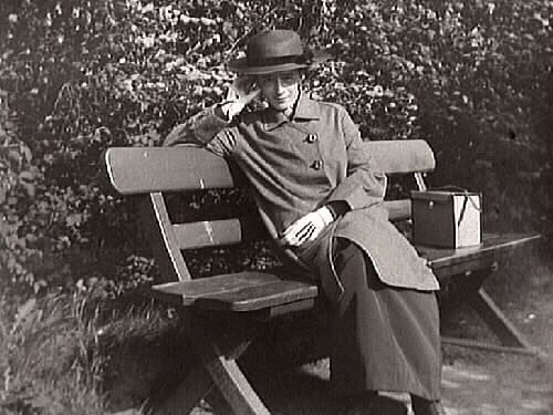 Fotografen fotograferar sig själv (?) utomhus vid häcken till grannfastigheten sittande på en bänk. Gården Vänhem, Tvååker. Foto från 1943-1944. Förvaringsbox till en kamera (?) bredvid henne på bänken.