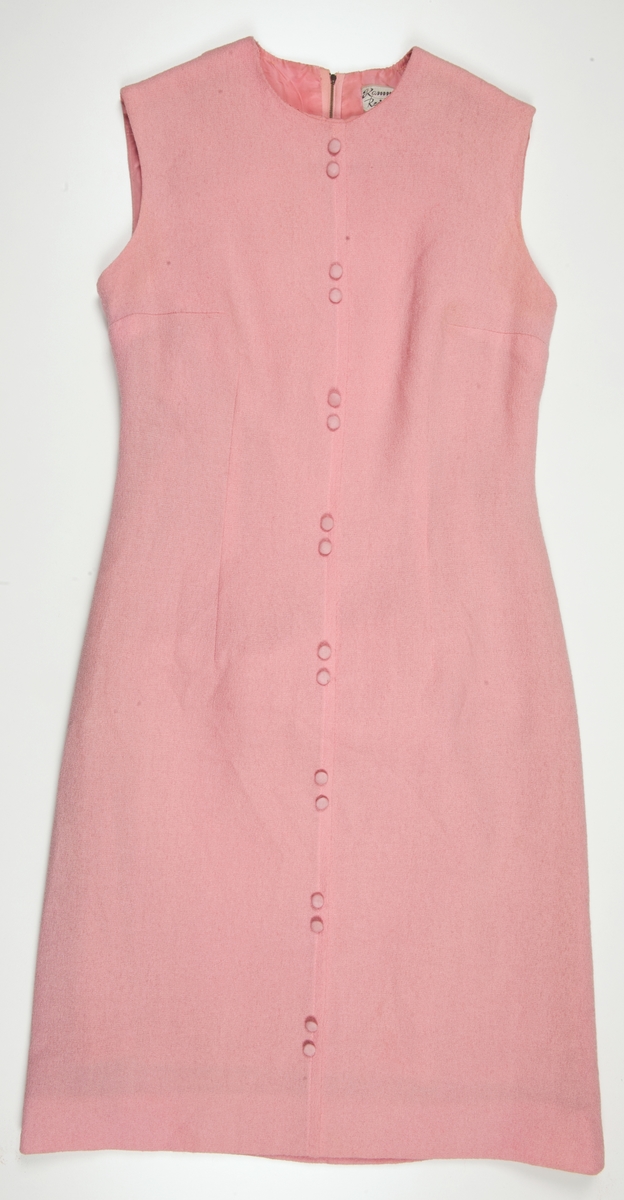 Ermeløs rosa kjole med rund hals og glidelås i ryggen. Små rosa stoffknapper på front. Fôr i rosa kunstsilke.