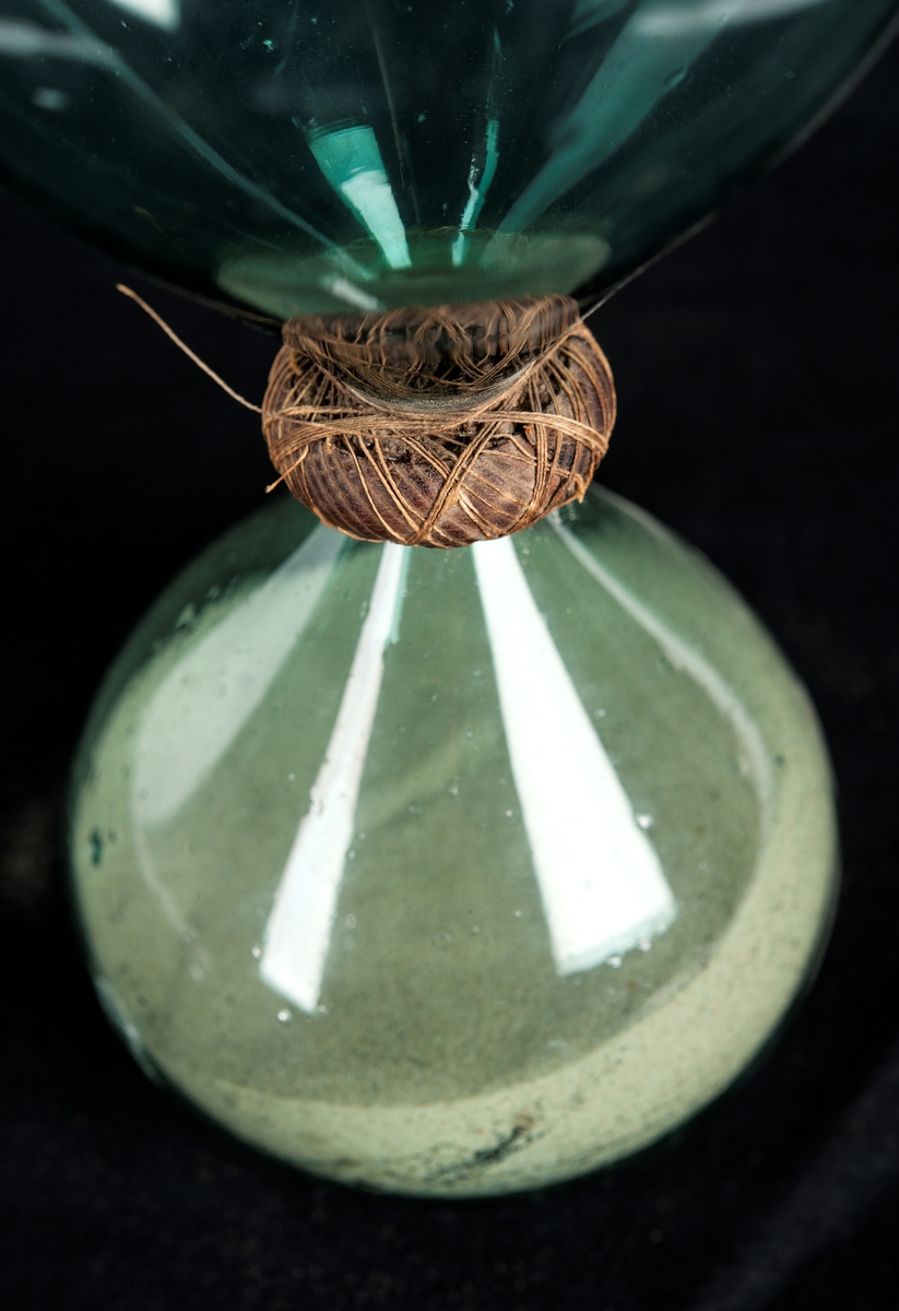Timglas hopsatt av två flaskor av ljusgrönt glas innehållande vit sand. Flaskorna är surrade med linnetråd i hopsättningen.