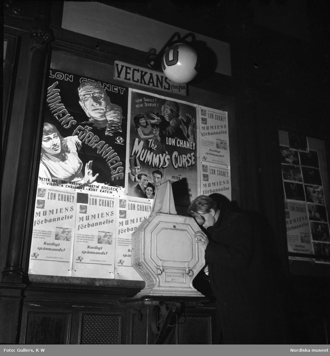 Eng ung man pressar ansiktet mot ett tittskåp utanför biograf vid Birger Jarlspassagen, Stockholm. Filmaffischer för "Mumiens förbannelse" (1944) på väggen.
