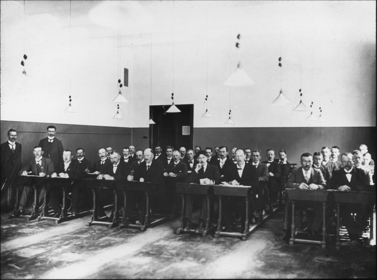 Bild visande skråverksamhet.
Gruppbild av män i lörosal, vid bänkar. Smeder, Göteborg, 1918.