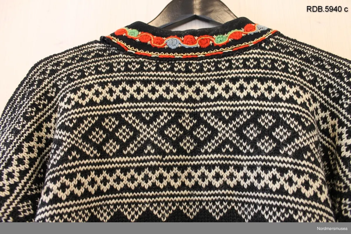 Håndstrikket Setesdalsgenser, herregenser, svart men kvitt mønster og med påsydd brodering i halsen. Broderiet har grønt fôr, og en tinnhekte påsydd foran. Genseren er slitt og hullet på albuene.