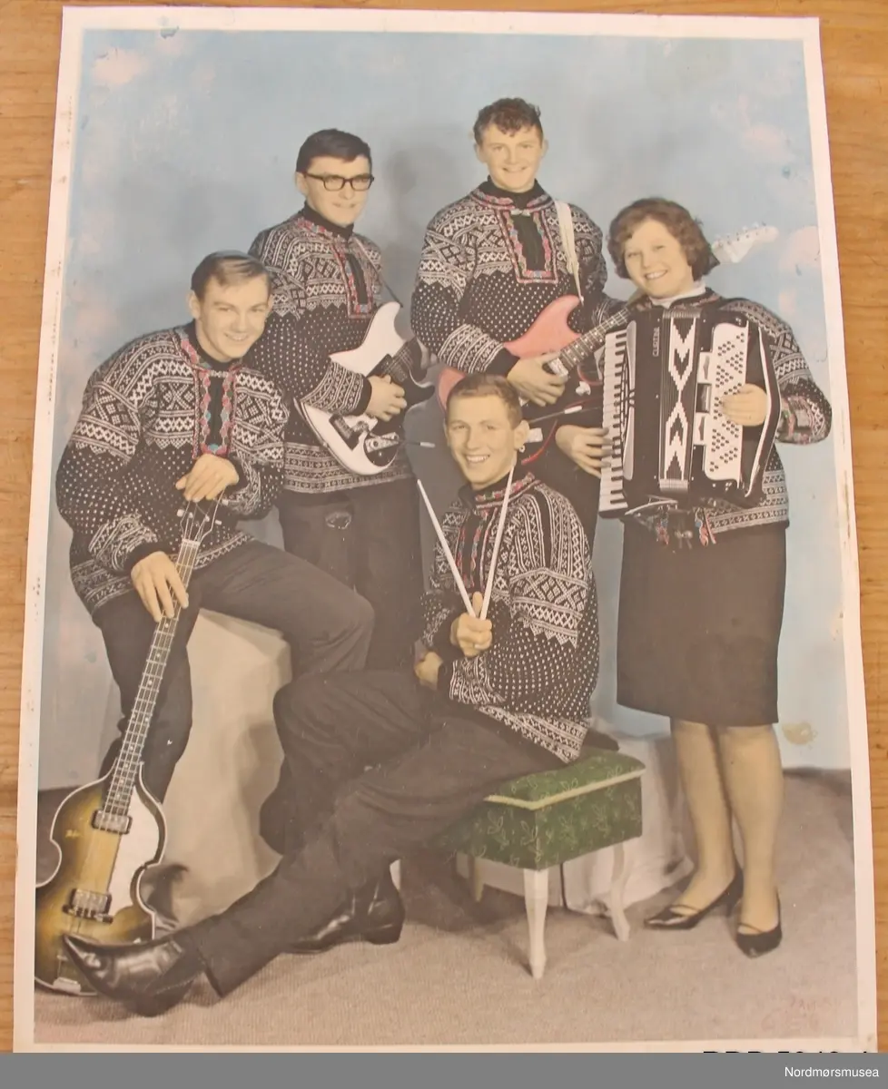 Håndstrikket Setesdalsgenser, herregenser, svart men kvitt mønster og med påsydd brodering i halsen. Broderiet har grønt fôr, og en tinnhekte påsydd foran. Genseren er slitt og hullet på albuene.