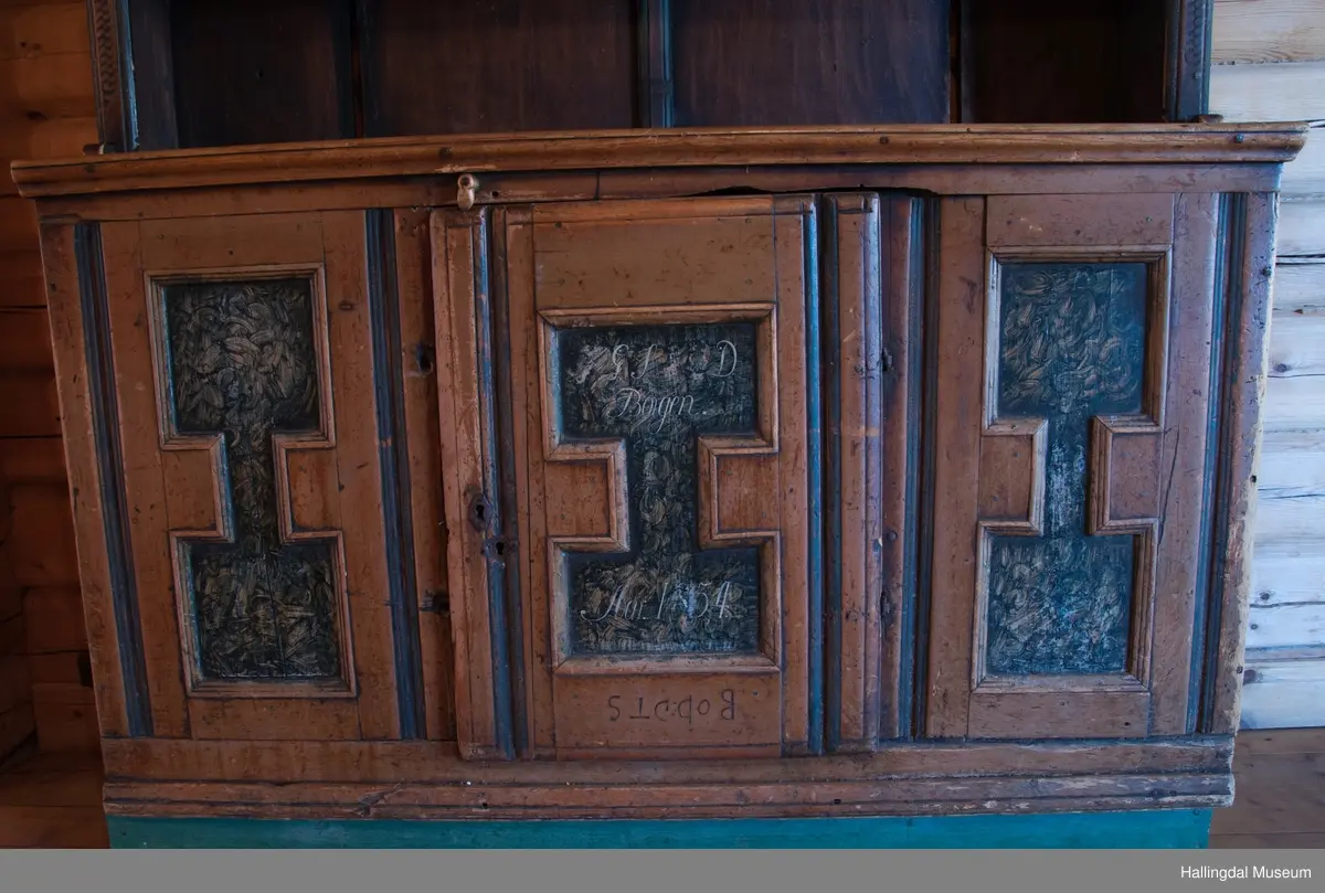 Rester etter rød og blå maling.  Gesims, skråner ned bakover. Profilert. 6 sirkulære hull i arkitraven.  Mellom disse er malt med sorte bokstaver.  Gavlene har utskårne "hylleknekter".  Bakveggen består av fire vertikale planker.  Skjøtene er dekket med profilerte lister.  Bare to biter i nederste del til venstre er fremdeles på plass.  Nederste del er lukket skap.  Tredelt, midterste del er skapdør.  Platen har profillist.  Feltene har speilfyllinger.  Blåmalte.  Klosset marmorering.  Speilfyllingenes form er som en liggen H.  Har profillist.  Skapet er satt på en ny grønmalt plate.  På døren er malt hvite bokstaver.  Nederst på døren er skåret inn bokstaver, opp-ned.  To nøkkelhull m. jernbeslag på venstre side av døren.  Over døren til venstre, en dreibar trekloss, holder igjen.  Innvendig har døren merke etter stort jernbeslag på samme side som nøkkelhullene.  To lister skåret i treverket minner om lange jernbeslag,-smalner mot nøkkelhullene.  Akantusranker i rødt, blått og hvitt, meget avslitt.  En hylle i skapet. Mangler bakvegg.  Karmen har på venstre side spor etter to jernhengsler.  Øverst på høyre kortside, mangler gesimsen en bit, og holdes til sideveggen med en jernkrampe.  Jernkrampe også på venstre side.  Skapplaten mangler list på høyre side.  Venstre side, fra gesimsen og ned har spor etter å ha stått inntil en vegg eller et annet møbel.