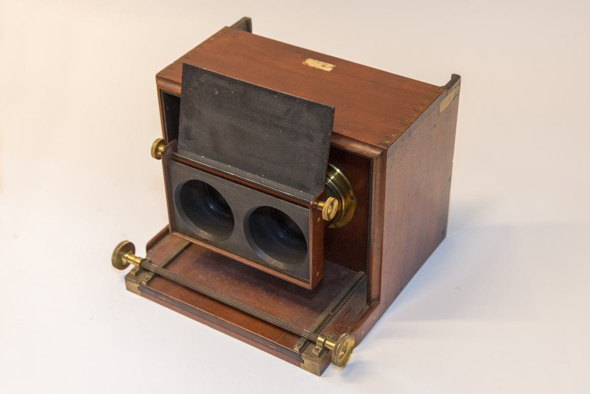 Gruppering med fotokamera i oppbevaringskasse, separat metall-lokk, stereolinse, lause delar som negativplate, mattskive, blendarsett og diverse smådelar.