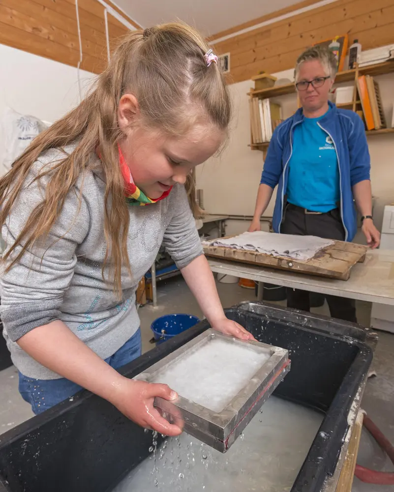 En Jente i skolealder står å lager papir, en dame står å følger med