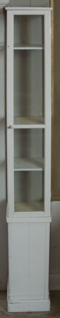 Monter av trä, vitmålad, stående rektangulär form. Glasad på tre sidor, dörr framtill med lås av vitmetall. Tre hyllplan av furu. Saknar bakstycke. Hög sockel.
