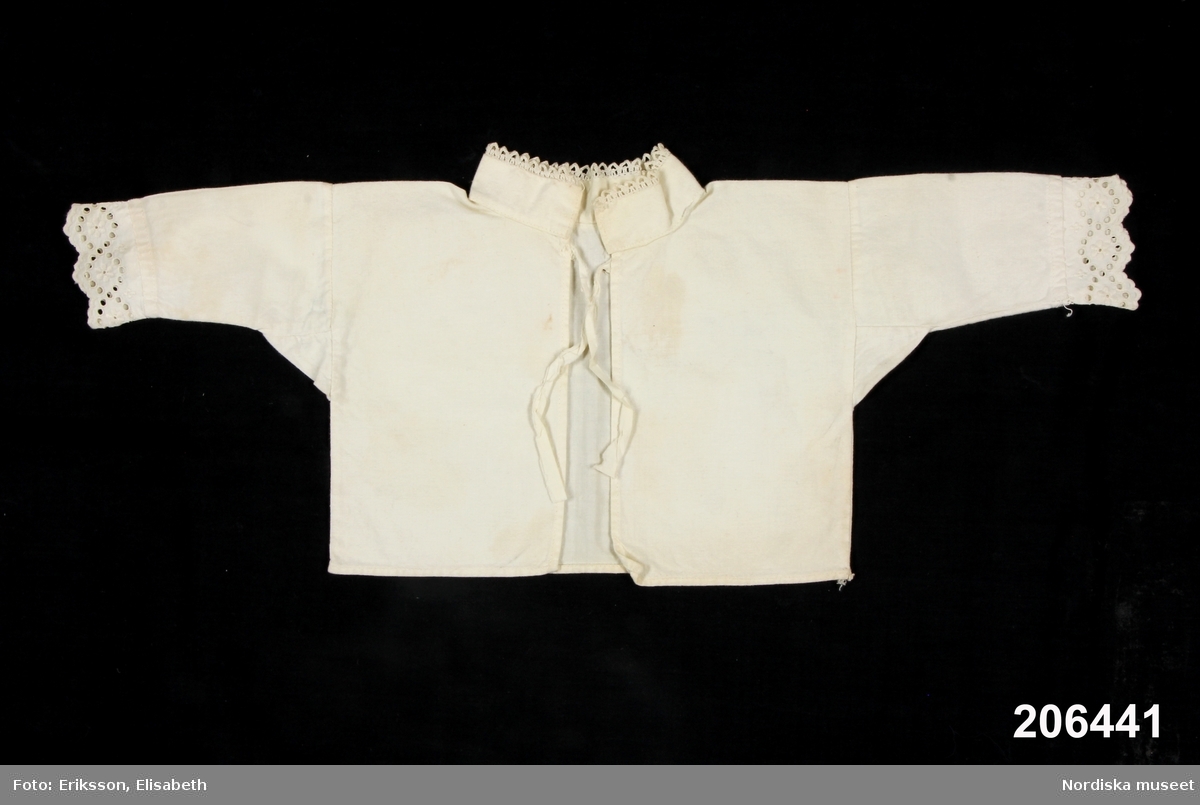 Spädbarnsskjorta av vit bomullslärft, ett bålstycke med sidsömmar, öppen fram med ett knytband i halsen, ståndkrage kantad med maskingjord uddspets. Vidsydd rak ärm med ärmsprjäll, i kanten maskingjord brodyr. Sydd på maskin.
Se även 206 442-446.
/Berit Eldvik 2013-02-07