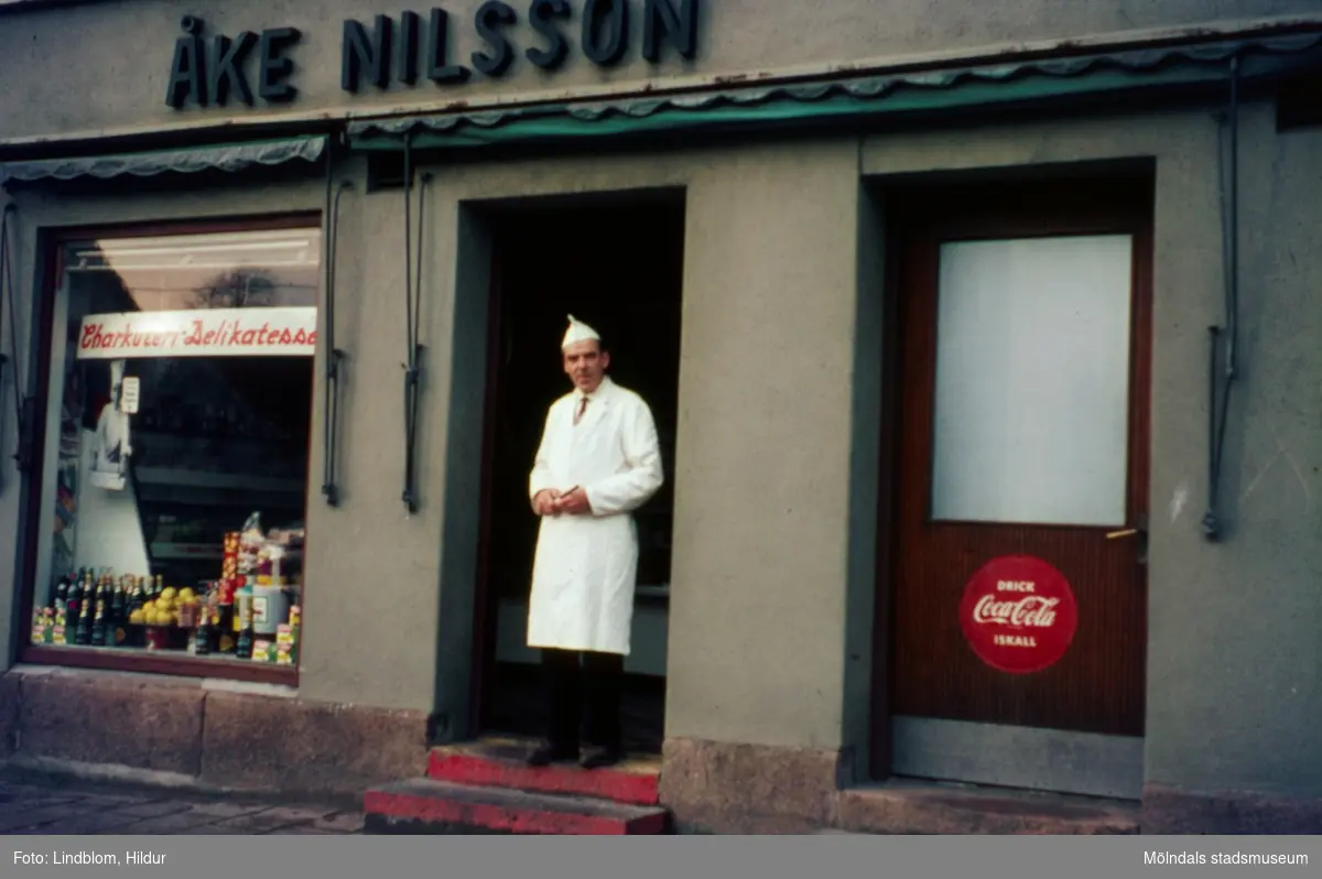 En man står i dörren till Åke Nilsson charkuteri och delikatesser med adress Kvarnbygatan 43 vid Gamla Torget i Mölndal, 1960-1970-tal.

För mer information om bilden se under tilläggsinformation.