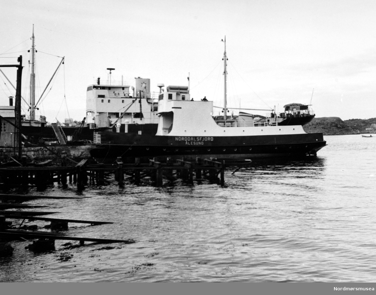 Bildet viser B/F";Norddalsfjord"; Storviks Mek. Verksted bnr. 14 ferdig utrustet ved piren. Båten vi ser bak ved piren er Ole T. Flakkes lastebåt M/S";Knoll";. "Norddalsfjord" ble levert til Møre og Romsdal Fylkesbåtar 15. mars 1961 og hadde følgende hoveddimensjoner: L 31,20 m x B 8,55 m x D 3,35 m og hadde en tonnasje på 159 brutto-registertonn. Fremdriftsmaskineriet består av 3 Volvo Penta turboladede dieselmotorer type TMD96 på til sammen 420 hk som via kilremdrift var koblet til et felles gir og propellaksel med vribar propell, slik at hver enkelt av motorene kunne kjøres separat. Fergen hadde 2 Bolinders vekselstrømsaggregater type 1052MG på 23 hk hver tilkoblet en generator på 17 kW. Fergen var utstyrt med elektrohydraulisk styremaskin. Fergen har plass til 18 personbiler og har sertifikat for 160 passasjerer. Forut er det innredet 6 lugarer for offiserer og restaurantpersonale og akterut en mannskapslugar for 4 personer og toppfarten er 11,4 knop og marsjfarten 10,5 knop. Ferga er verkstedets første nybygg etter B/F"Trygge" som ble levert i 1938. I forgrunnen ses bakenden på den nye klinkbygde patentslippvogna i stål fra 1918, den største slippen mellom Bergen og Trondheim, som ble erstattet med nytt slippanlegg ca. 1979, og banene til såpehellingen fra 1901 nærmest. Bygget helt til venstre inneholder en dampkjel og nederst i høyre hjørne ses litt av trekaia nedenfor støperiet. Bak ferga ses Teistholmen på Gomalandet. Bildet er fra 1961. Tekst av Peter Storvik. Fra Nordmøre museums fotosamlinger.



