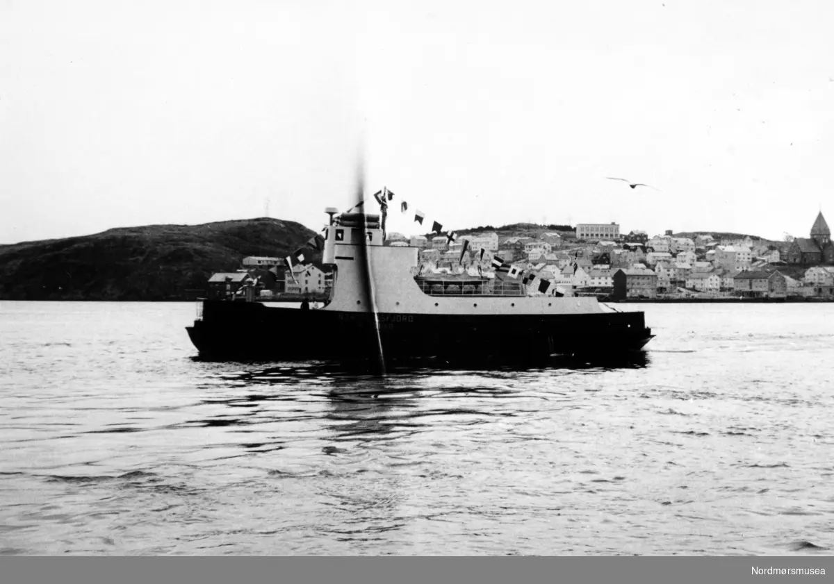 Bildet viser B/F"Norddalsfjord" Storviks Mek. Verksteds bnr.14 på overleveringsturen på havna i Kristiansund. Vi ser at signalflaggene, rederiflagget og det norske flagget er heist.

"Norddalsfjord" ble levert til Møre og Romsdal Fylkesbåtar 15. mars 1961 og hadde følgende hoveddimensjoner: L 31,20 m x B 8,55 m x D 3,35 m og hadde en tonnasje på 159 bruttoregistertonn. Fremdriftsmaskineriet består av 3 Volvo Penta turboladede dieselmotorer type TMD96 på til sammen 420 hk som via kilremdrift var koblet til et felles gir og propellaksel med vribar propell, slik at hver enkelt av motorene kunne kjøres separat. Fergen hadde 2 Bolinders vekselstrømsaggregater type 1052MG på 23 hk hver tilkoblet en generator på 17 kW. Fergen var utstyrt med elektrohydraulisk styremaskin.

Fergen har plass til 18 personbiler og har sertifikat for 160 passasjerer. Forut er det innredet 6 lugarer for offiserer og restaurantpersonale og akterut en mannskapslugar for 4 personer og toppfarten er 11,4 knop og marsjfarten 10,5 knop.

Ferga er verkstedets første nybygg etter B/F"Trygge" som ble levert i 1938.

Personene på dekk og på brua er vanskelige å kjenne igjen.

I bakgunnen ses Nordlandet og Bjørnehaugen.

Bildet er fra 1961. Kilde/tekst: Peter Storvik. Fra Nordmøre museums fotosamlinger.
