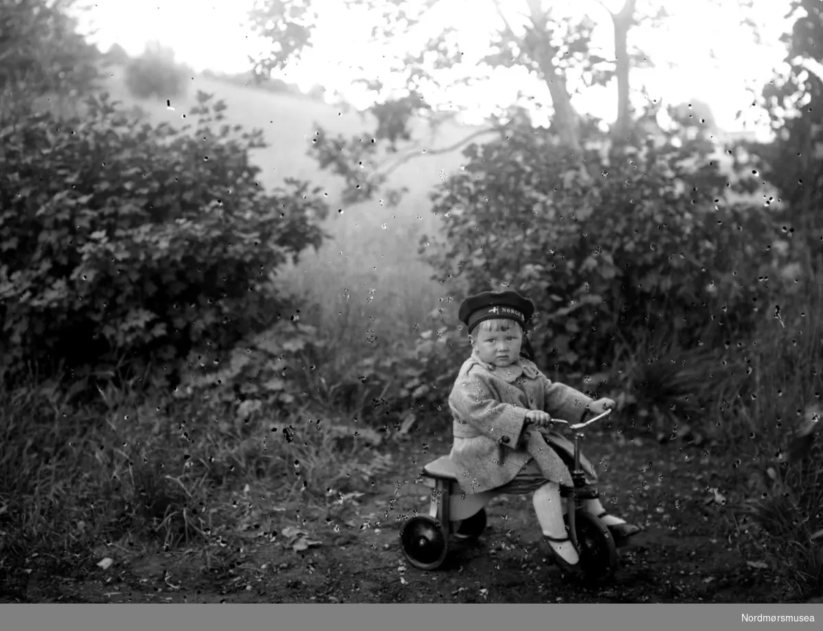 Foto av et ukjent barn på trejulsykkel en plass på Nordmøre, Dette er ett av barnebarna til Margrethe og Georg Sverdrup..
 Ukjent datering. Dublett av KMb-1987-005.4145. Fra Nordmøre Museums fotosamlinger.

