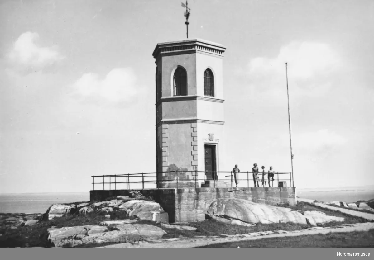 Postkort av Vardetårnet på Kirkelandet i Kristiansund, set fra sør. Her ser vi diverse barn og voksne som står langs platten. Gammel vaktplass 78 m.o.h. med storslagen utsikt til skipsleia med Grip i horisonten. Panoramaet går 360 oC rundt med Nordmørsfjellene som mektig bakgrunn. Et gammelt tretårn ble i 1892, til byens 150-års jubileum, erstattet med t karakteristisk murtårn. Dette tårnet ble revet i 1976 og erstattet med en kopi som ble innviet 8 mai 1983. I 1992 ble en kopi av det gamle tretårnet gitt i gave til Kristiansund kommune fra utflyttede kristiansundere i Oslo.

Opprinnelig har dette punktet 78 m.o.h fungert som varde og vaktstue for den optiske telegrafen mellom Kristiansund og Trondheim i 1808. Herfra er det flott utsikt til skipsleia med Grip i horisonten. Denne vaktplassen spilte en viktig rolle da byen ble angrepet av to engelske fregatter i 1808. Senere ble det brukt av tollvakten, før det i 1849 ble solgt til seilmaker Andreas Cornelius Hansen.

I perioden 1840 til 1880 sto tårnet åpent for alle og ble bl.a. hyppig brukt til kortspill og dans. Mange besøkende fant veien hit da som nå, og disse har etterlatt sine merker på veggene; årstall og navn, initialer m.m. fantes på alle de seks originale veggene, deriblant flere spanske navn.

Vardetårnet i mur ble reist for å kaste glans over byens 150 års jubileum i 1892. Vardetårnet erstattet dermed det langt mindre og enklere tretårnet, som sto på samme sted.

Under første verdenskrig beslaga marinen tårnet og brukte det som vakthus. Senere ble det brukt som salgsbod for datidens godterier. Varden var et kjært samlingspunkt for familier på søndagsutflukt. Lærer Meegaard sørget for å plassere en langkikkert sammen med et kart som viste omgivelsene. Under 2. verdenskrig bygde tyskerne tak over øverste etasje, og det ble da benyttet som vakttårn.

Etter krigen ble vedlikeholdet av tårnet nedprioritert, og det endte med at Vardetårnet fra 1892 ble revet i mai 1976 da det etter sigende var ";farlig for omgivelsene";. Kommunen hadde, på grunn av anstrengt økonomi, ikke planer om å reise et nytt tårn. Etter privat initiativ og innsamlede midler ble et nytt tårn finansiert og bygget av ildsjeler i ";Vardens venner";. Det nye tårnet ble høytidelig åpnet 8. mai 1983 med en enorm folkemengde tilstede.

I 1992 ble også en tro kopi av det gamle sekskantete tretårnet gitt som gave til Kristiansund kommune fra utflyttede kristiansundere i Oslo, etter initiativ av Arne Sunde. Det ble laftet på Hjerleidske videregående skole på Dombås.

Opprinnelig stod dette tårnet på samme sted som Vardetårnet står i dag. Det er tre meter høyt, og taket er formet som et klippfiskflak. Opp gjennom sentrum av taket rager en jernstang som har vært brukt som vindretningsviser. I dag står det originale tårnet ved Norsk klippfiskmuseum, og både kompassrosetten på innsiden av taket og de mange skribleriene på veggen, er godt bevart


(kilde: http://www.kristiansund.no/artikkel.aspx?MId1=1619&amp;AId=2179&amp;Back=1 , som refererer til Ove Borøchstein: ";En bauta ble reist"; 2003 Kristiansund kommune";).            
Fra Nordmøre Museums fotosamlinger. Reg: EFR
