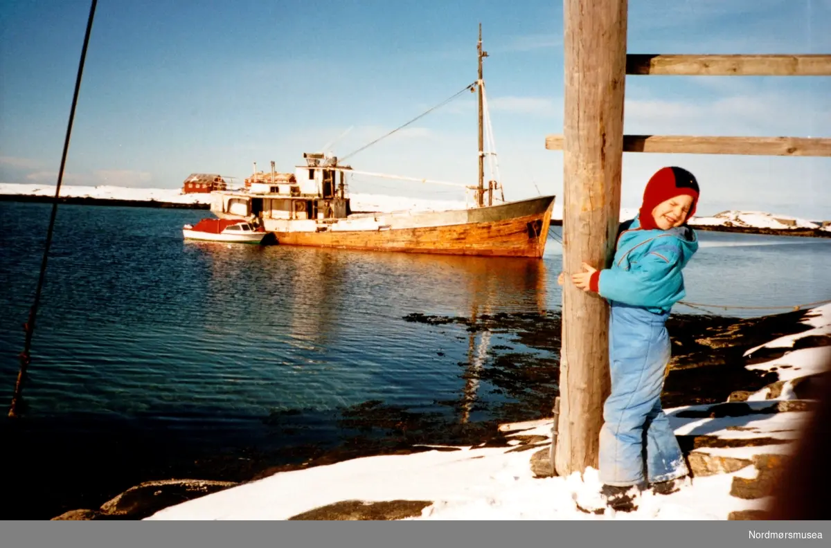 Foto av en gutt stående ved en trepåle med en fiskebåt liggende ute i fjorden like bak. Det er ukjent hvem vi ser på bildet, samt hvor og når bildet er tatt. Trolig er det fra Møre og Romsdal en plass (Averøy? Kristiansund?) omkring 1980 til 1999. Fra Nordmøre museums fotosamlinger. Reg: EFR
