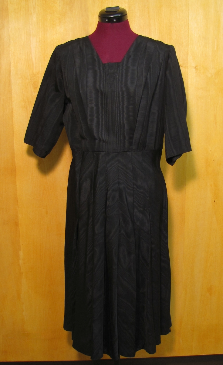 Klänning i svar moiré som var modetyget på 1940-talet. Klänning sydd till givaren Berta Ågrens syster. Klänningen har halvlång ärm och på kjolen nedsydda veck.