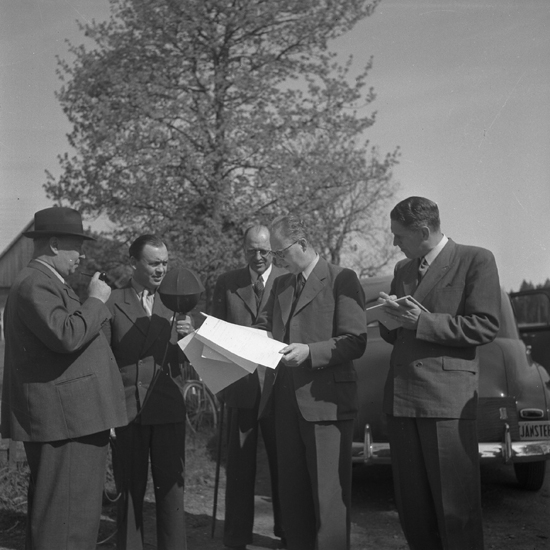 En man intervjuar några män medan en av dem håller i ett papper och besvarar frågor. Till höger står en man 
och antecknar på ett block. En äldre man röker pipa.