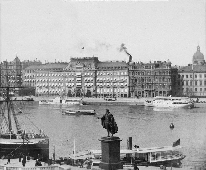 Foto på en kaj med båtar. I bakgrunden står det hus på rad. På ett av husen är det en skorsten som ryker. Vid hamnen står en staty.
