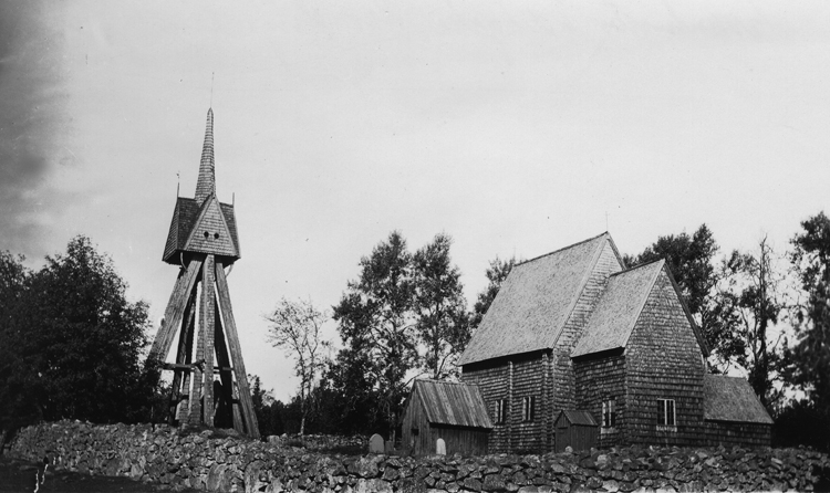 Foto en klockstapel och en liten kyrka med en stenmur utanför.