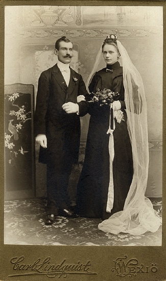 Porträtt av ett brudpar (helfigur). Hon är klädd i svart brudklänning med hög krage och vit slöja med myrtenkrona. Han är iklädd frack och vita handskar m.m.
Till vänster om sig har brudgummen en skärm med japaninspirerat motiv. I bakgrunden syns en kuliss med jugendmönster m.m.

Kan ev. vara David Natanael Olofsson (1870-1957), Växjö, m. fru. 
Källa: Sveriges Dödbok 1947-2003.