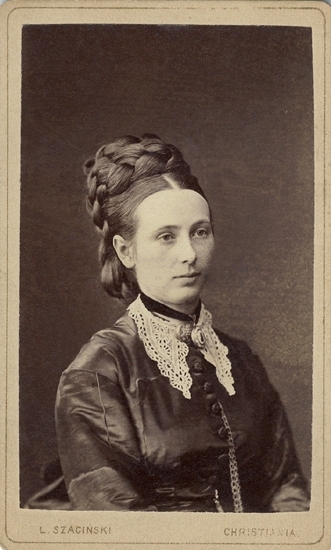 Porträtt (bröstbild, halvprofil) av en kvinna i flätfrisyr och mörk sidenklänning med spetskrage. Runt halsen har hon ett sammetsband med en medaljong. Dessutom skymtar en brosch i halsgropen. På bröstet syns också en klockkedja. 


Fru Trine Aubert - kan ev. vara Anna Cathrine Sommerfeldt (1849-1887),
g. 1867 i Christiania (nuv. Oslo) med  Ludvig Mariboe Benjamin Aubert, (1838-  ) , sedan 1864 professor i juridik vid universitetet i Christiania. 
Källa: Internet.