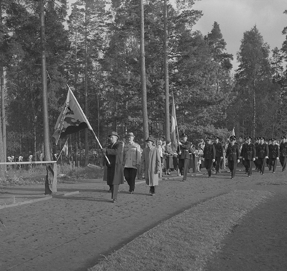 Svenska Flaggans Dag, 6/6 1953. 
Några herrar i rock och hatt tågar in på Värendsvallen bärande Kungl. Kronobergs regementes kamratförenings fana, ,
följda av poliser i uniform.