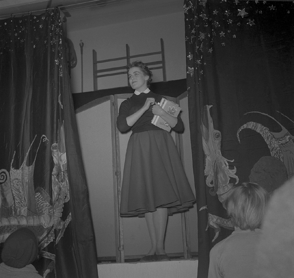 Sjuornas fest, 16/2-17/2 1954.
En flicka i kjol och blus m.m. talar om något, med en
bunt böcker i famnen.