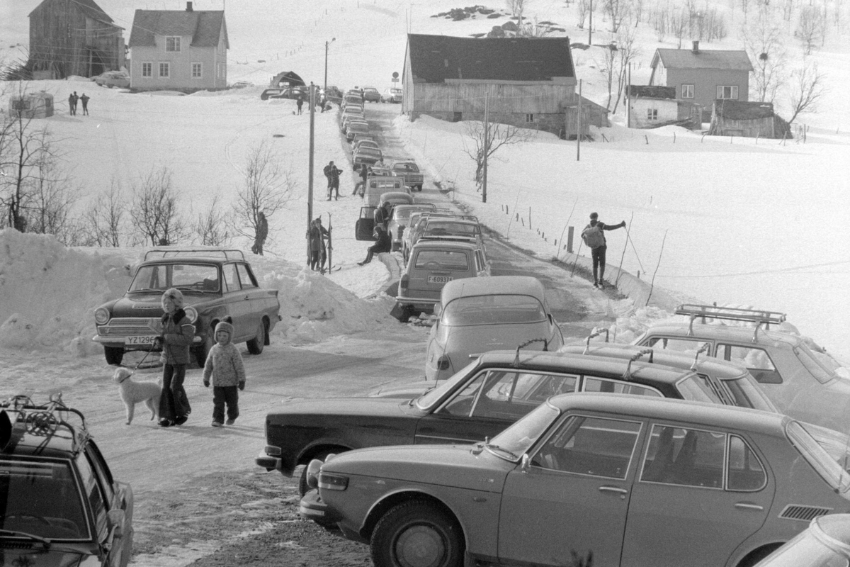 Parkerte biler og skiløpere i Sørlia. Bebyggelse i bakgrunnen.