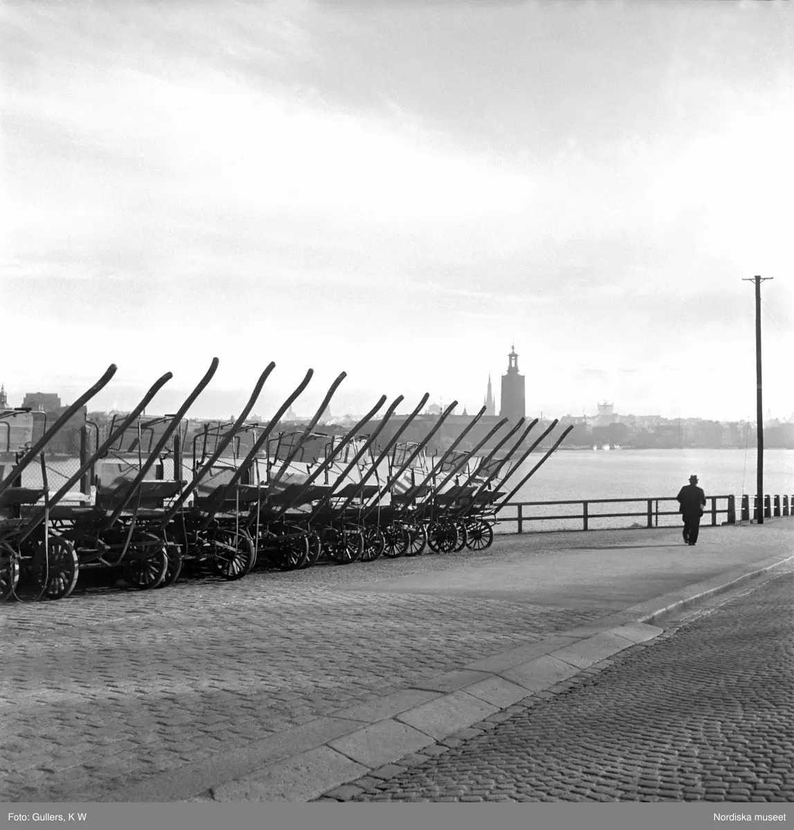 Stockholm, Söder Mälarstrand. Hästkärror uppställda på rad längs kajkanten. Riddarfjärden och Stadshuset i bakgrunden.