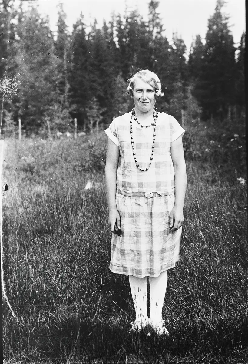 Ung kvinne stående i rutet kjole i en eng. Dette er Inga Haug f. Inga Moen