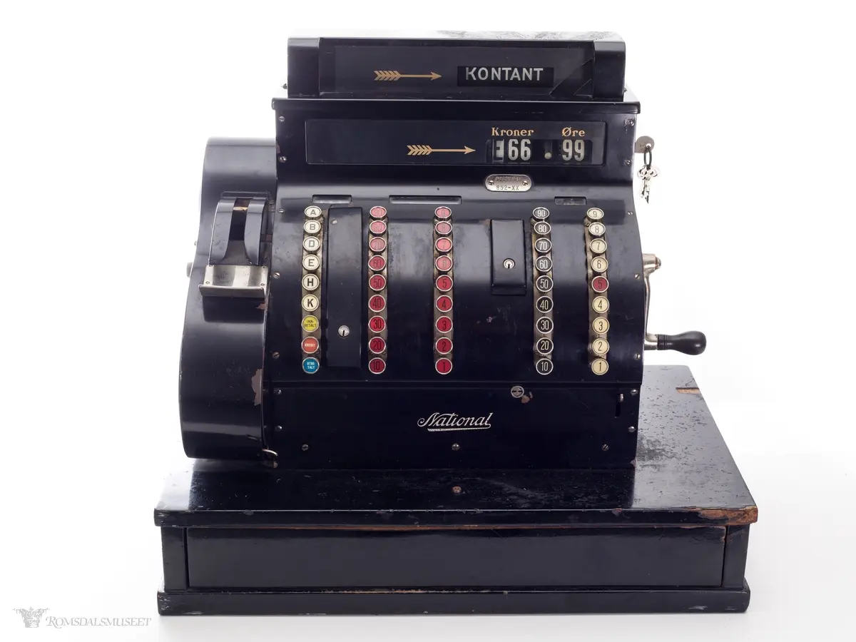 Sortfarget kassaapparat av merke National. Et kontantfelt øverst på apparatet med et felt under for tresifret kronebeløp og tosifret øre. Tre mindre felter over taster merket "KKK", "Kroner" og "Øre".

Fem rader med knapper i front. 
Raden lengst til venstre har bokstaver A-K, samt knapper for "innbetaling", "kreditt" og "utbetaling"
To tasterader på venste side med tall fra 1-9 og 10-90, og tilsvarende på høyre side.
Boks for utskrift av kvittering på vesntre side(?)

Sveiv og nøkler i lås på høyre side og av og på skru på venste side.