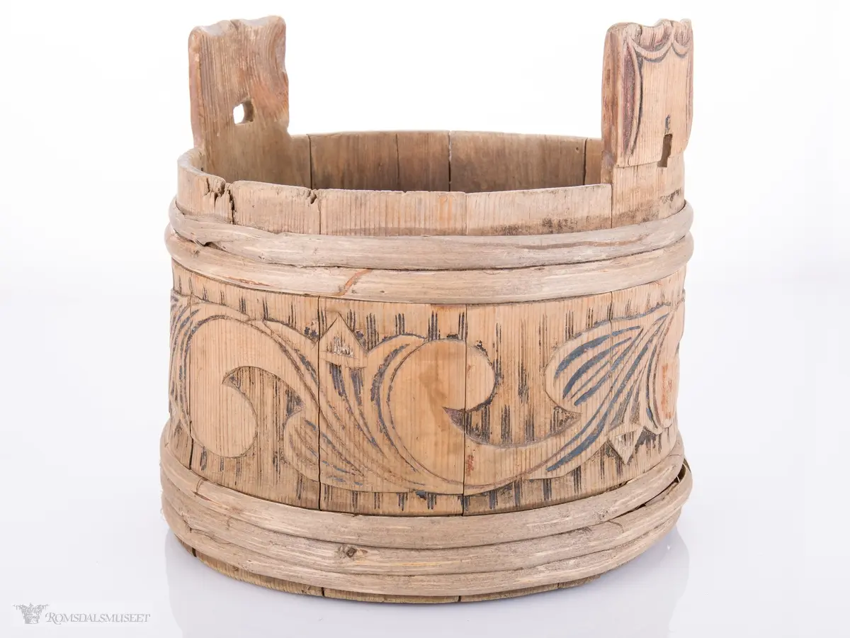 Rund med 3 vidjeband nede og 2 oppe og ører til lokk som mangler. Utskjæringer i relieff på hver stav.