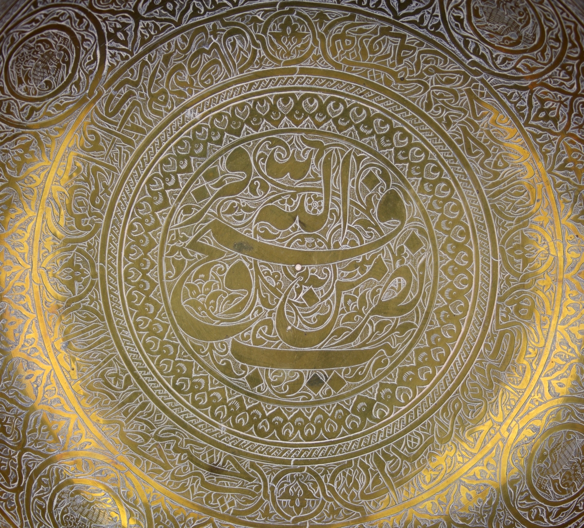 Skål av mässing med graverad dekor.Enligt notering från Persien. På skålen finns både arabisk och persisk skrift. Det är citat ur koranen (Med hjälp av gud är segern nära) och en persisk dikt.
