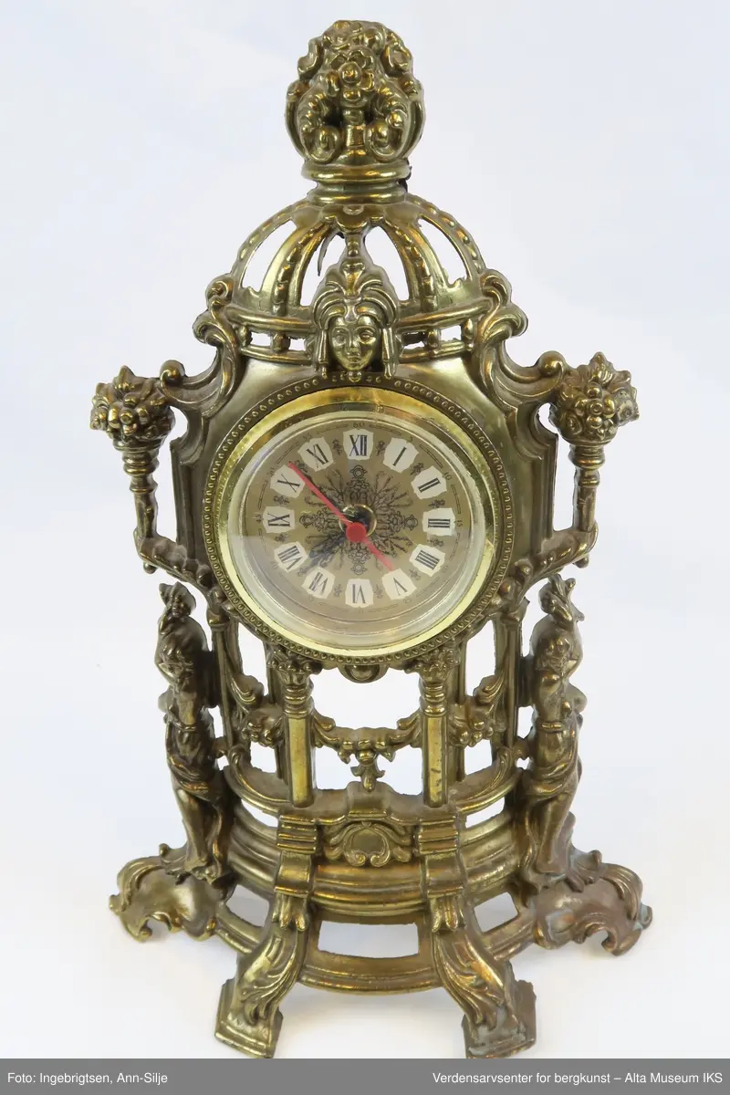 Batteridrevet ur. Stående oppsats med barokk innfatning og innfelt urskive med romertall.