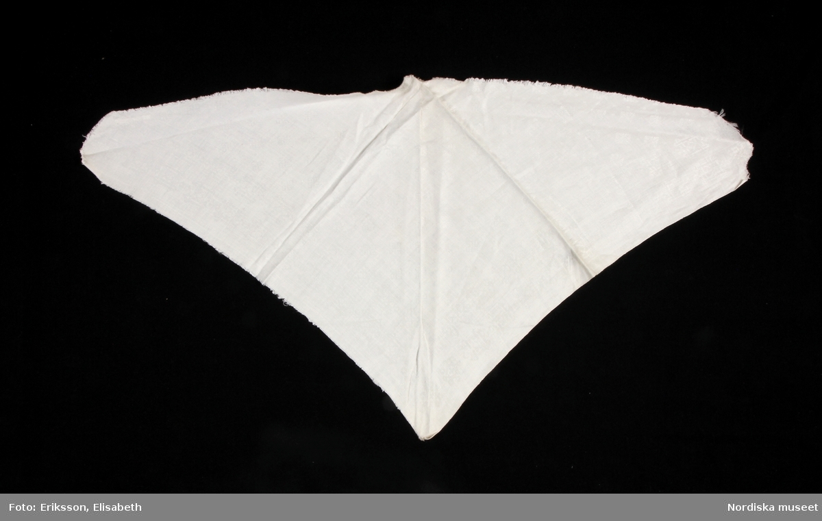 Halskläde i vit damast med fornnordiskt mönster. Tresnibb med stadkant på ena kortsidan, andra kortsida och långsida klippt kant.