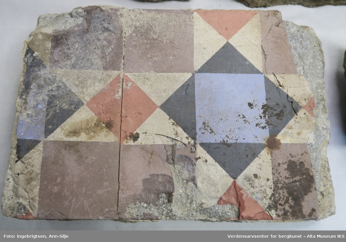 Bruddstykker av gulvfliser med dekor av geometriske fargefelter med tre- og firkanter. Flisene er støpt fast i sement.